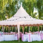Festlich dekorierter rosa Pavillon mit runden Tischen und Stuehlen, umgeben von Lichterketten im gruenen Garten
