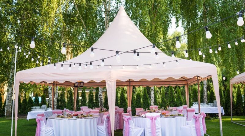 Festlich dekorierter rosa Pavillon mit runden Tischen und Stuehlen, umgeben von Lichterketten im gruenen Garten